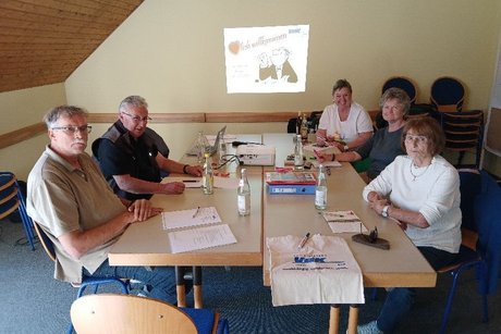 Seminarraum Klausur mit 5 Personen um einen Tisch mit Getränken und Beamer. Bild vom Beamer mit Willkommen.