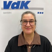 Auf dem Bild ist eine Frau mit zusammengebundenen Haaren, Brille, braunem Kragen und schwarzem Pulli zu sehen.