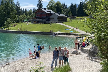 Am Achensee drei Personen, ein Mann und zwei Frauen der Ortsgruppe VdK Garching und mehrere Badegäste am Achensee, im Hintergrund die Gaisalm