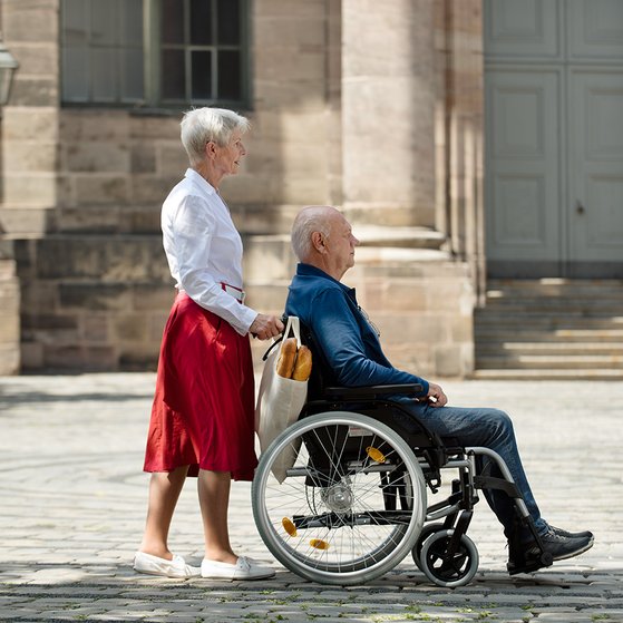 Eine Seniorin schiebt ihren Mann, der im Rollstuhl sitzt, &uuml;ber einen Platz
