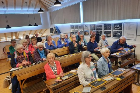 Die Teilnehmenden sitzen in einem alten Klassenzimmer und hören die Ausführungen der Museumsführerin.