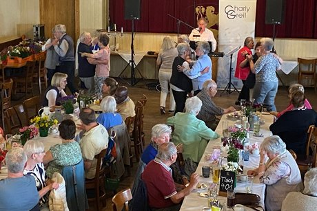 mehrere Personen sitzen an l&auml;nglichen Tischen mit wei&szlig;en Tischdecken und Blumenschmuck mit Getr&auml;nken. Die Menschen h&ouml;ren der Musik zu oder unterhalten sich untereinander.Bildes sitzt der Musiker, davor sind einige Tanzpaare, die zu der Musik tanzen