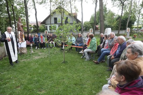 Das Bild zeigt Pater Dan Anzorge in seinem Garten des Lebens mit den Teilnehmern an der Maiandacht bei herrlichem Wetter im Schatten der hochgewachsenen Bäume