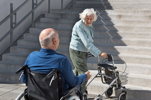Ein &auml;lterer Mann im Rollstuhl und eine &auml;ltere Frau mit Rollator stehen am Fu&szlig; einer un&uuml;berwindbaren Treppe.