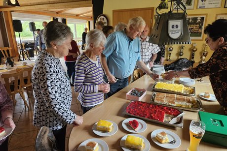 auf einem Tisch stehen drei Kuchenbleche und einige Teller mit Kuchen einige G&auml;ste. 5 Personen warten auf den Kuchen, den die Kassiererin verteilt