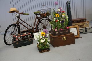 Bild zeigt ein altes Fahrrad, alte verschiedene alte Koffer auf dennen Blumen stehen;