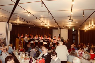 Menschen sitzen im Rathaussaal, Auf der Bühne im Hintergrund steht der Singkreis