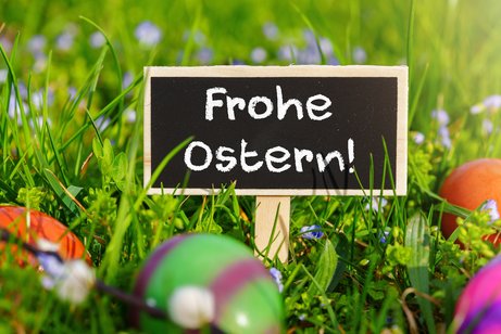 Kleines schwarzes Tafelschild mit der Aufschrift &bdquo;Frohe Ostern!&ldquo; im Fr&uuml;hlingsgras, umgeben von bunten Ostereiern und kleinen Blumen.