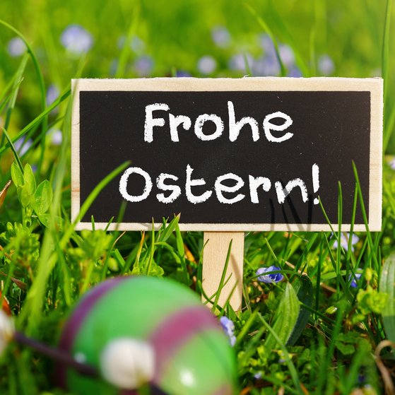 Kleines schwarzes Tafelschild mit der Aufschrift &bdquo;Frohe Ostern!&ldquo; im Fr&uuml;hlingsgras, umgeben von bunten Ostereiern und kleinen Blumen.