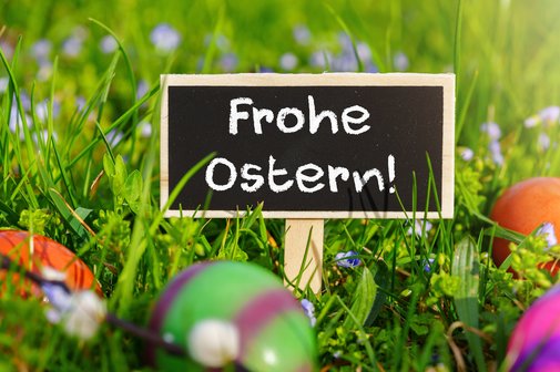 Kleines schwarzes Tafelschild mit der Aufschrift &bdquo;Frohe Ostern!&ldquo; im Fr&uuml;hlingsgras, umgeben von bunten Ostereiern und kleinen Blumen.