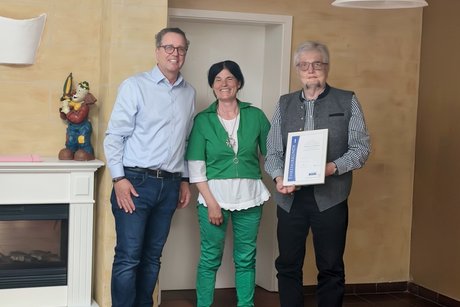 Das Bild zeigt drei Personen die Kamera l&auml;cheln. Links steht ein Mann, der erste B&uuml;rgermeister von Heroldsbach, mit hellblauem Hemd und dunkler Jeans. In der Mitte steht eine Frau, die erste Vorsitzende des VdK Ortsverbandes in gr&uuml;ner Jacke und Hose mit wei&szlig;er Bluse und wei&szlig;en Schuhen. Rechts steht ein Herr mit Brille, dunkler Weste und kariertem Hemd und h&auml;lt eine gerahmte Urkunde in den H&auml;nden. Die W&auml;nde sind in warmen Beige- und Braunt&ouml;nen. Die Stimmung wirkt feierlich, es handelt es sich um eine Ehrung f&uuml;r 50 Jahre im VdK Bayern.