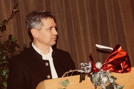 Bürgermeister Weihnachtsgruss Bürgermeister steht auf Bühne hinter Rednerpult. Im Vordergrund Weihnachtsdeko mit rotem Weihnachtsstern und Efeu und Weihnachtsschmuck in Silber