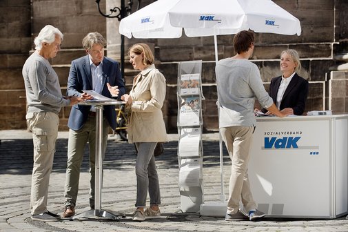 Ein VdK-Infostand in einer Fu&szlig;g&auml;ngerzone, eine Frau spricht mit einem Passanten, am Stehtisch daneben unterhalten sich Menschen. 