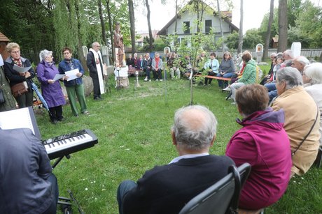 Das Bild zeigt Pater Dan Anzorge in seinem Garten des Lebens mit den Teilnehmern an der Maiandacht bei herrlichem Wetter im Schatten der hochgewachsenen Bäume