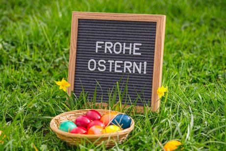 Holzschild mit der Aufschrift &bdquo;Frohe Ostern!&ldquo; steht im gr&uuml;nen Gras, davor ein kleiner Korb mit bunt gef&auml;rbten Ostereiern und gelbe Narzissen.