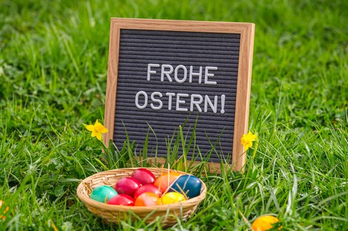 Holzschild mit der Aufschrift &bdquo;Frohe Ostern!&ldquo; steht im gr&uuml;nen Gras, davor ein kleiner Korb mit bunt gef&auml;rbten Ostereiern und gelbe Narzissen.