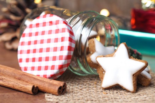 Auf einem Tisch liegt ein Weckglas, das mit Zimtsternen gefüllt ist. Links daneben liegen drei Zimtstangen. 