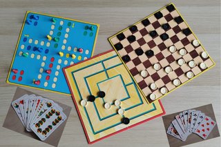 Bild zeigt drei Brettspiele mit Spielsteinen (Mensch ärger dich nicht, Damespielbrett und Mühlenspielbrett) und zwei Kartenspielblätter Skat und Schafkopf auf einem Tisch.