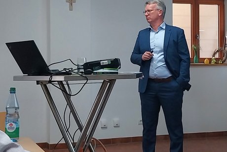 Referent steht am Stehtisch, darauf liegt ein Notebook, auf welches er blickt.