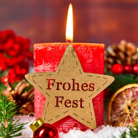 Eine brennende rote Kerze, verziert mit einem Stern mit der Aufschrift: "Frohes Fest". Umgeben von weihnachtlicher Dekoration.
