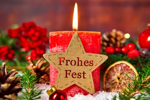 Eine brennende rote Kerze, verziert mit einem Stern mit der Aufschrift: "Frohes Fest". Umgeben von weihnachtlicher Dekoration.