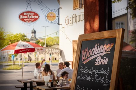 Auf dem Bild sieht man die Außenansicht der Gastwirtschaft Aubräu Traunstein - im Hintergrund erblickt man die über 400 Jahre alte Salinenkirche in der Au - die größte Kapelle Deutschlands. Im Vordergrund ist ein Werbeaufsteller mit den Tagesangeboten und den Essensschmankerl. Dahinter sitzen Gäste an Tischen bei einer Brotzeit und Bier. Auf dem Bild sieht man auch ein Zunftzeichen der Brauerei und der Gastwirtschaft.