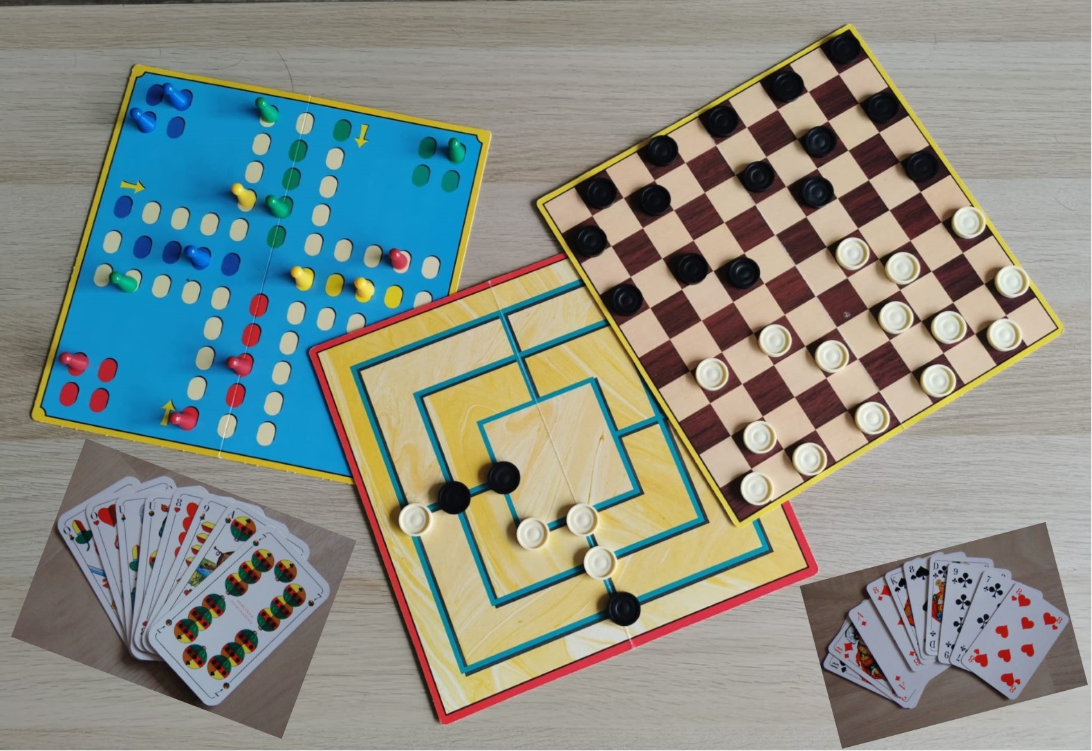 Bild zeigt drei Brettspiele mit Spielsteinen (Mensch ärger dich nicht, Damespielbrett und Mühlenspielbrett) und zwei Kartenspielblätter Skat und Schafkopf auf einem Tisch