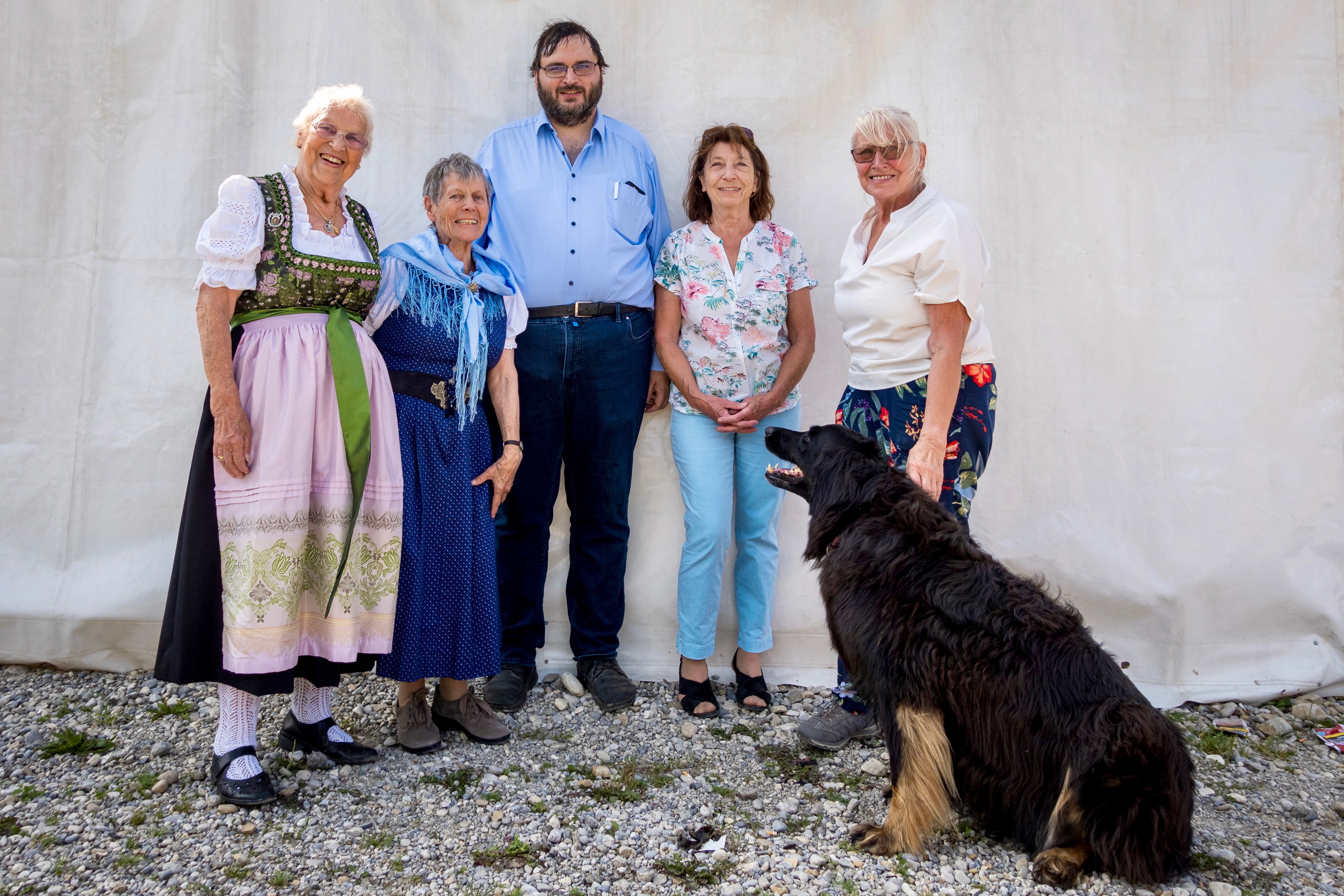 Das Bild zeigt die Vorstandschaft des VdK Ortsverbands Aubing vor einem hellen, neutralen Hintergrund. Fünf Personen stehen nebeneinander und blicken lächelnd in die Kamera. Von links nach rechts sind zu sehen: Helga Brandl in einem traditionellen Dirndl mit rosa Schürze, Wilma Frömmel in einem blauen Kleid mit hellblauem Schal, Ludwig Bauer mit Brille in einem hellblauen Hemd, Johanna Moll in einer weißen Bluse mit Blumenmuster und Ingrid Steger mit heller Kurzhaarfrisur in einer weißen Bluse und bunter Hose. Vor der Gruppe sitzt die Hündin Ema, ein großer, langhaariger schwarzer Hund, der freundlich in Richtung der Menschen schaut.
