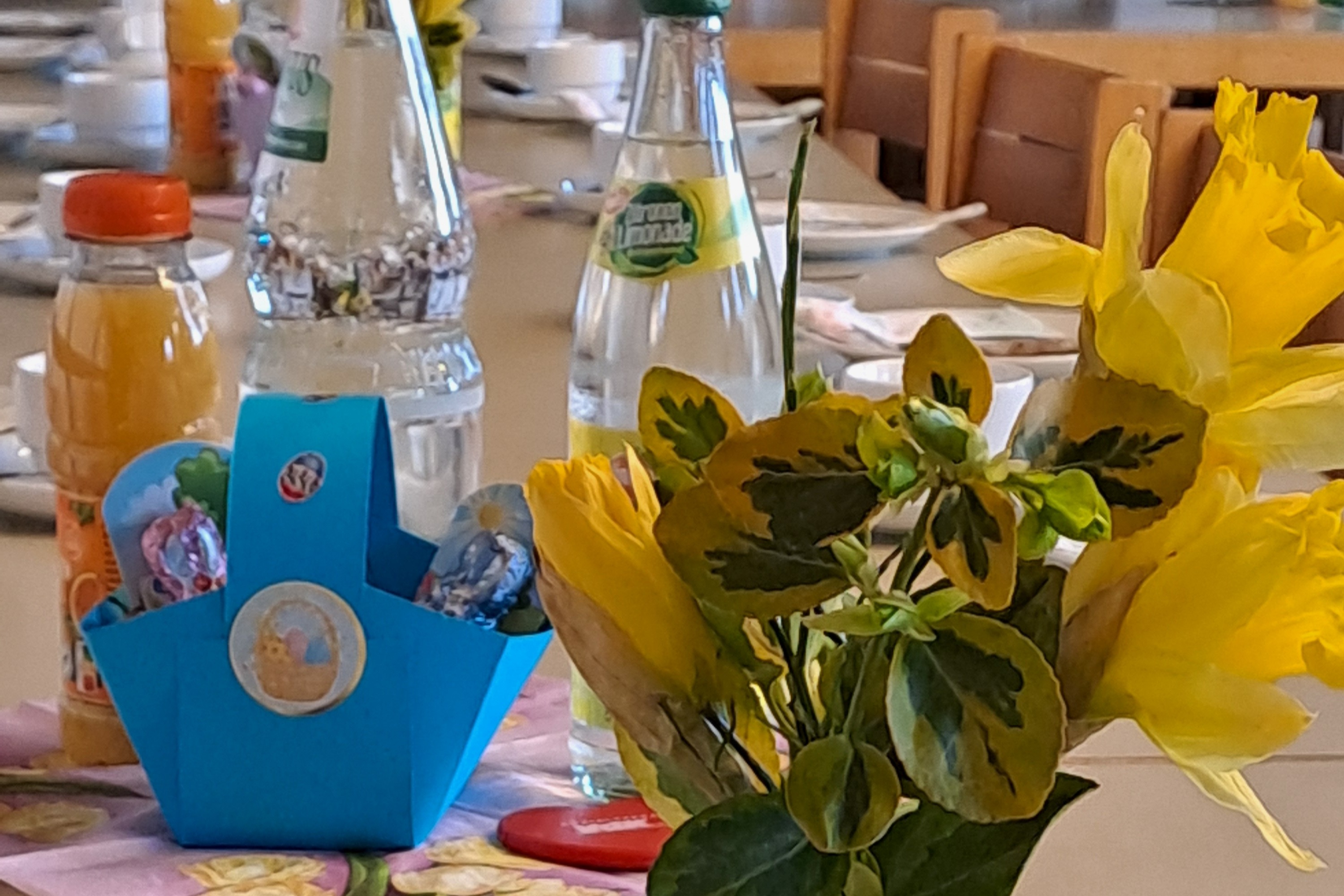 Auf dem Bild sieht man einen Tisch eine Vase mit einem Strau&szlig; Osterglocken, dahinter ein Osterk&ouml;rbchen in blau und Getr&auml;nke.