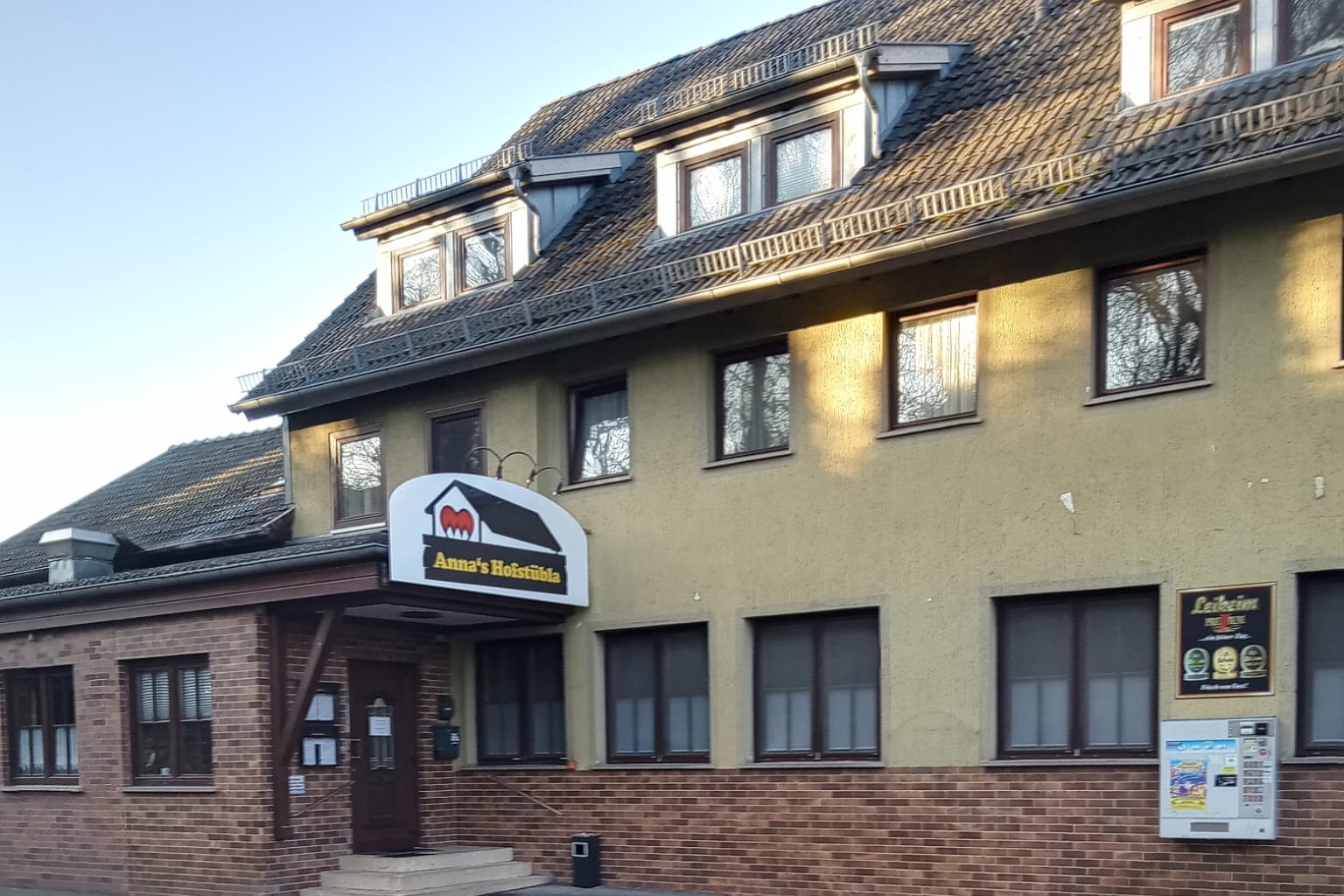 Stra&szlig;enansicht des Haupteingeng zum Gasthaus &bdquo;Anna&rsquo;s Hofst&uuml;bla mit Werbetafel und Tabackautomat