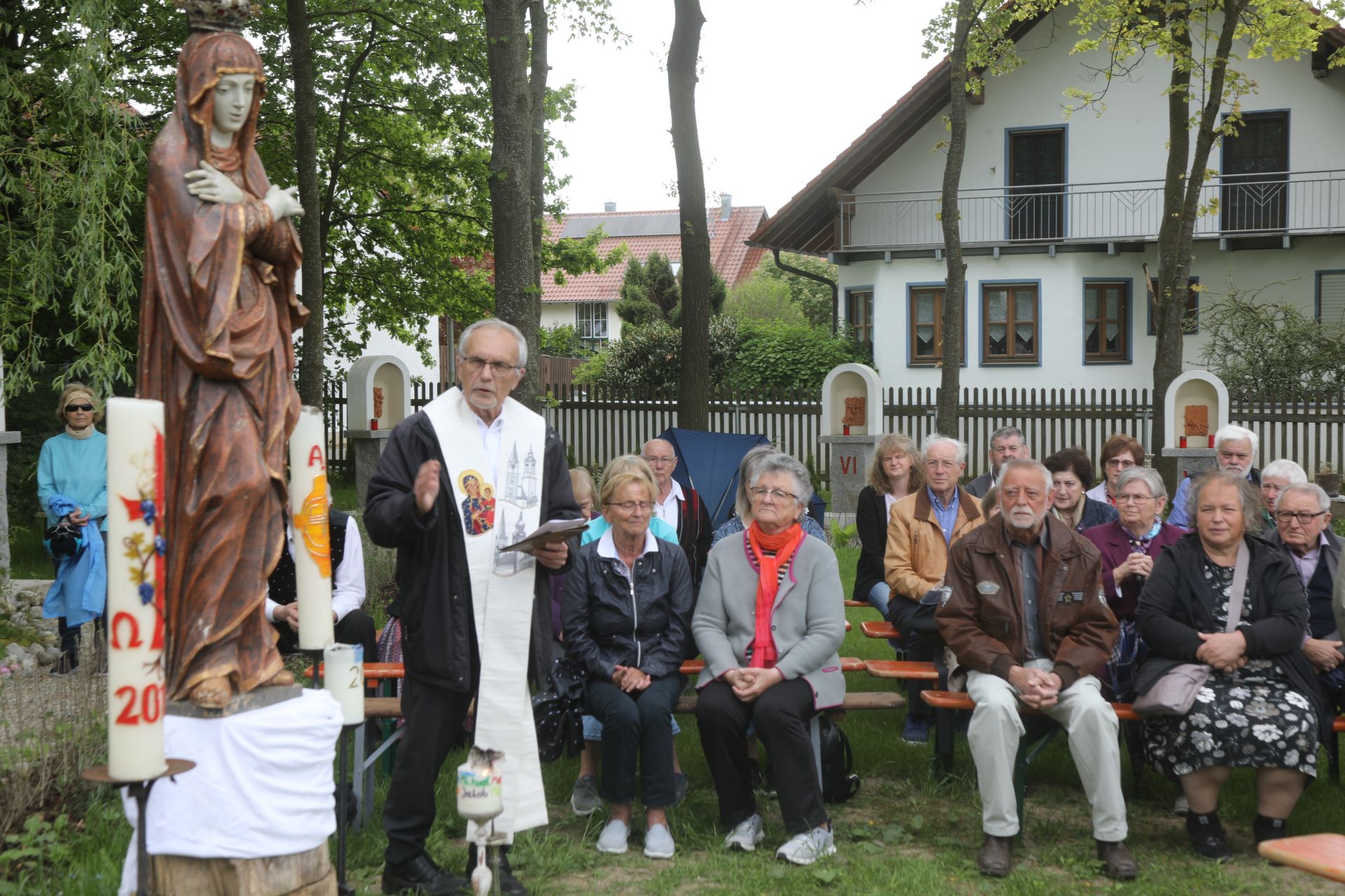 Das Bild zeigt Pater Dan Anzorge in seinem Garten des Lebens mit den Teilnehmern an der Maiandacht bei herrlichem Wetter im Schatten der hochgewachsenen Bäume