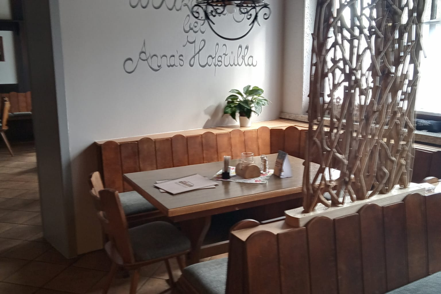 Blick in den vorderen Gastraum, an der Wand "Willkommen in Anna' Hofst&uuml;bla, U-F&ouml;rmige Eckbank mit G&auml;stetisch, Raumteiler mit Pflanzen, Fenster zur Hauptstra&szlig;e