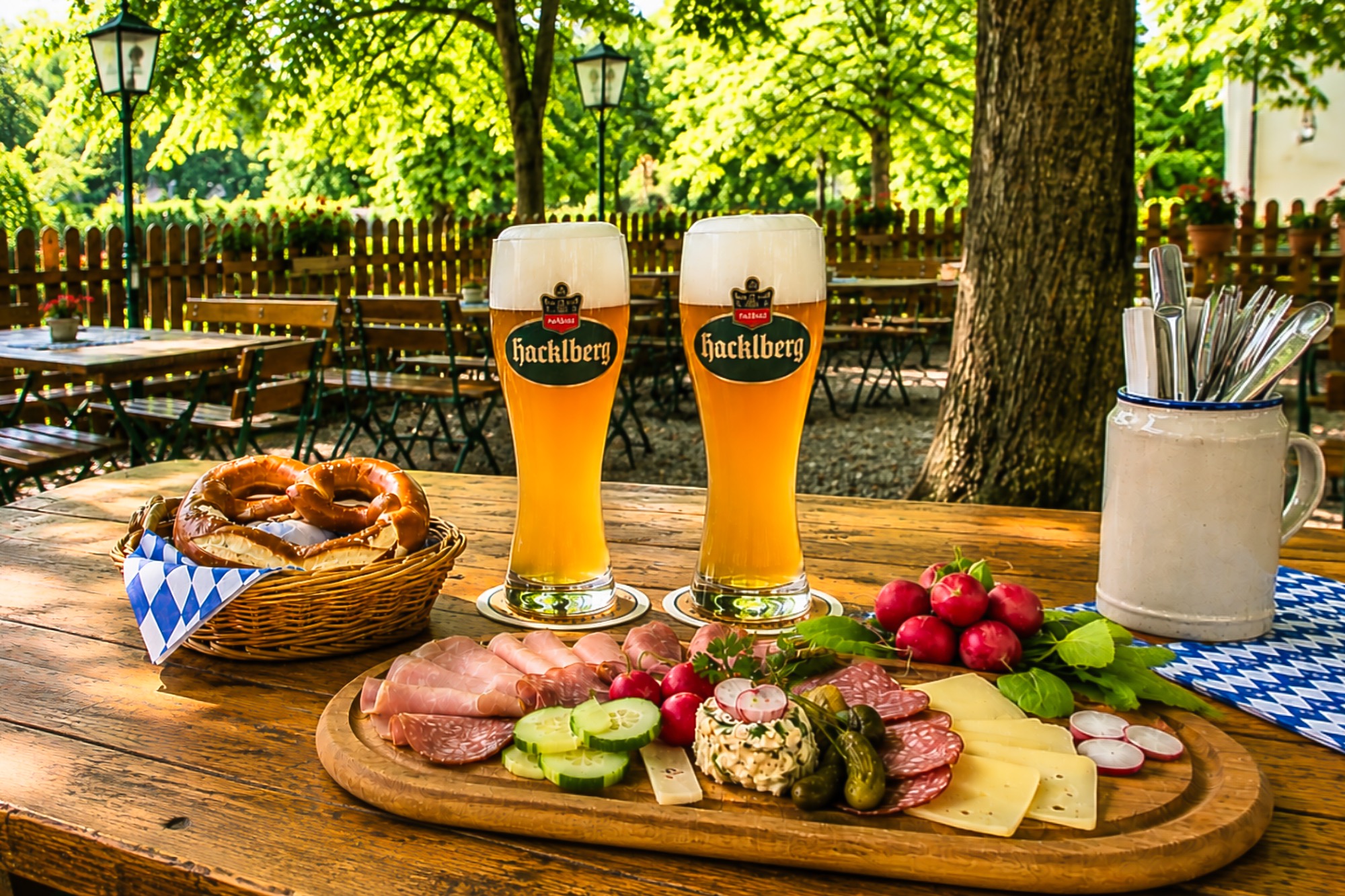 Brotzeit im Biergarten, auf dem Tisch stehen 2 Wei&szlig;biere und ein Brotzeitteller