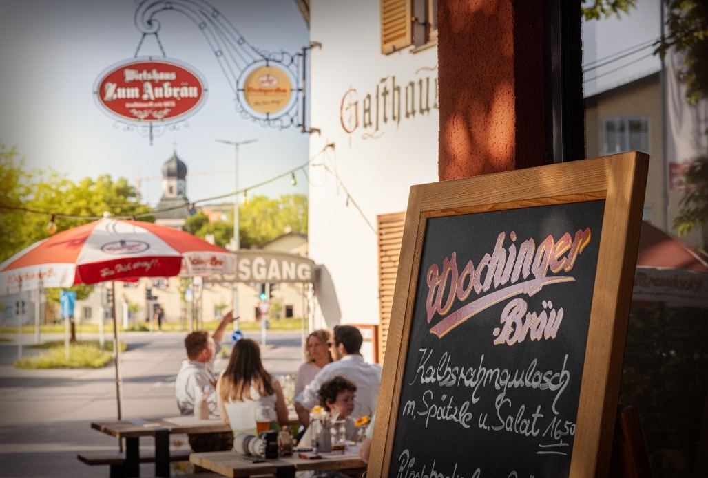 Auf dem Bild sieht man die Außenansicht der Gastwirtschaft Aubräu Traunstein - im Hintergrund erblickt man die über 400 Jahre alte Salinenkirche in der Au - die größte Kapelle Deutschlands. Im Vordergrund ist ein Werbeaufsteller mit den Tagesangeboten und den Essensschmankerl. Dahinter sitzen Gäste an Tischen bei einer Brotzeit und Bier. Auf dem Bild sieht man auch ein Zunftzeichen der Brauerei und der Gastwirtschaft.