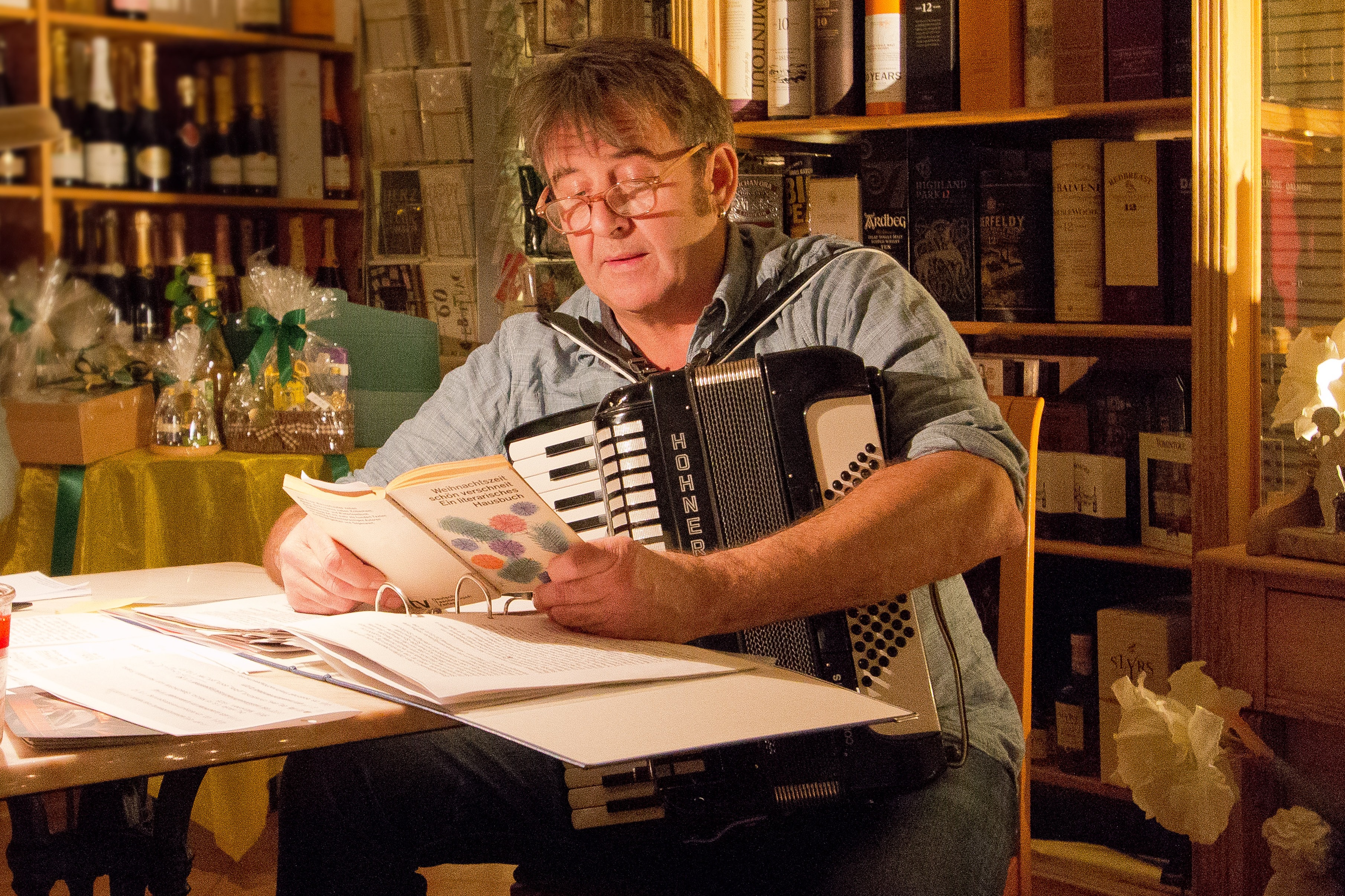 lesender Herr Ritsch Ermeier am Tisch, Buch in der Hand, auf dem Tisch vor ihm ein aufgeschlagener Ordner in einem Zimmer einer Buch- und Weinhandlung