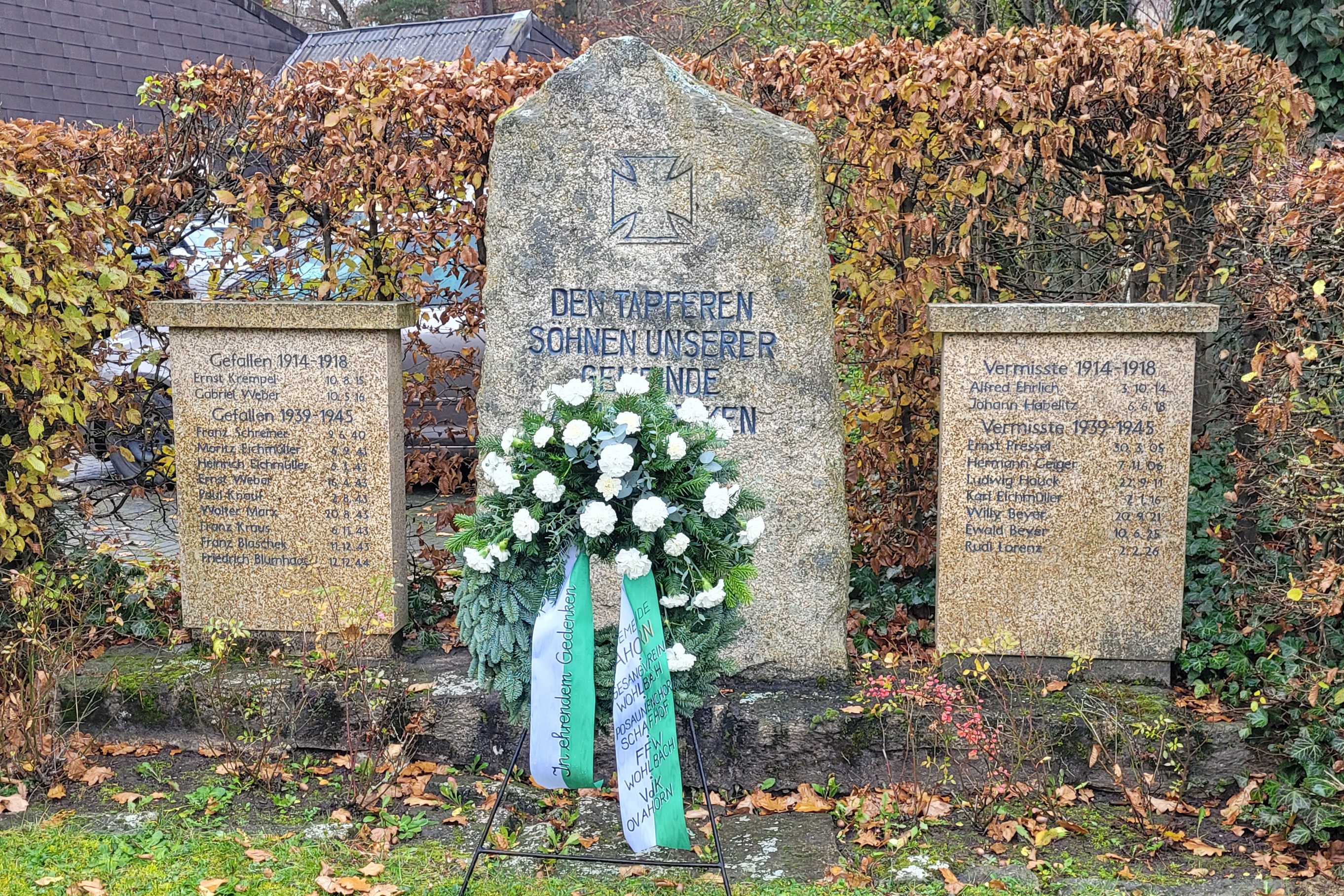Ehrenmal vor einer Hecke Gedenkstein in der Mitte rechts und links die Namentafel aus Stein