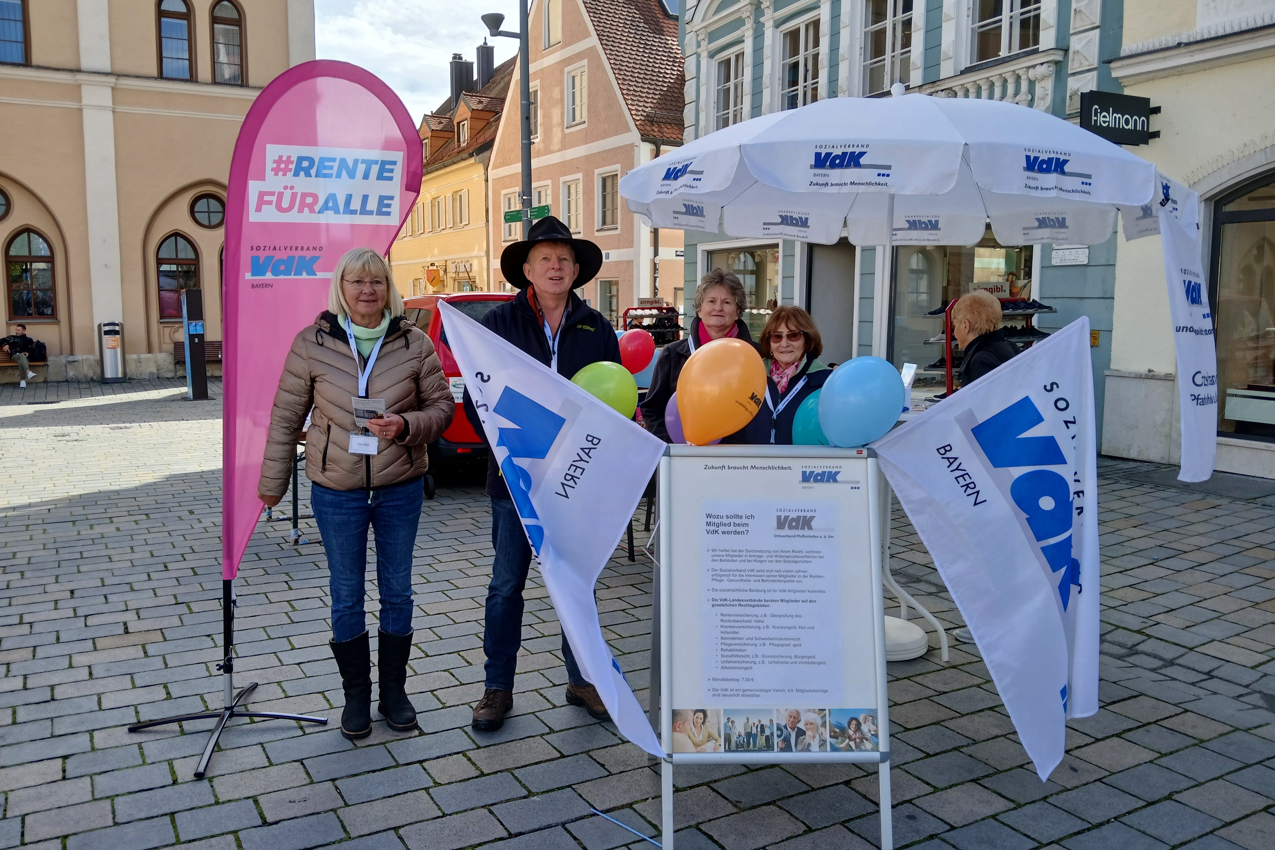 VdK Informationsstand: Infoständer mit Luftballon und 2 VdK-Fahnen recht und links. VdK-Sonnenschirm mit Tisch und Informationsmaterialien, rosa Stoffschild mit Aufschrift: Rente für alle und 3 Frauen und ein Mannin einer sonnigen Passage der Innenstadt Pfaffenhofens
