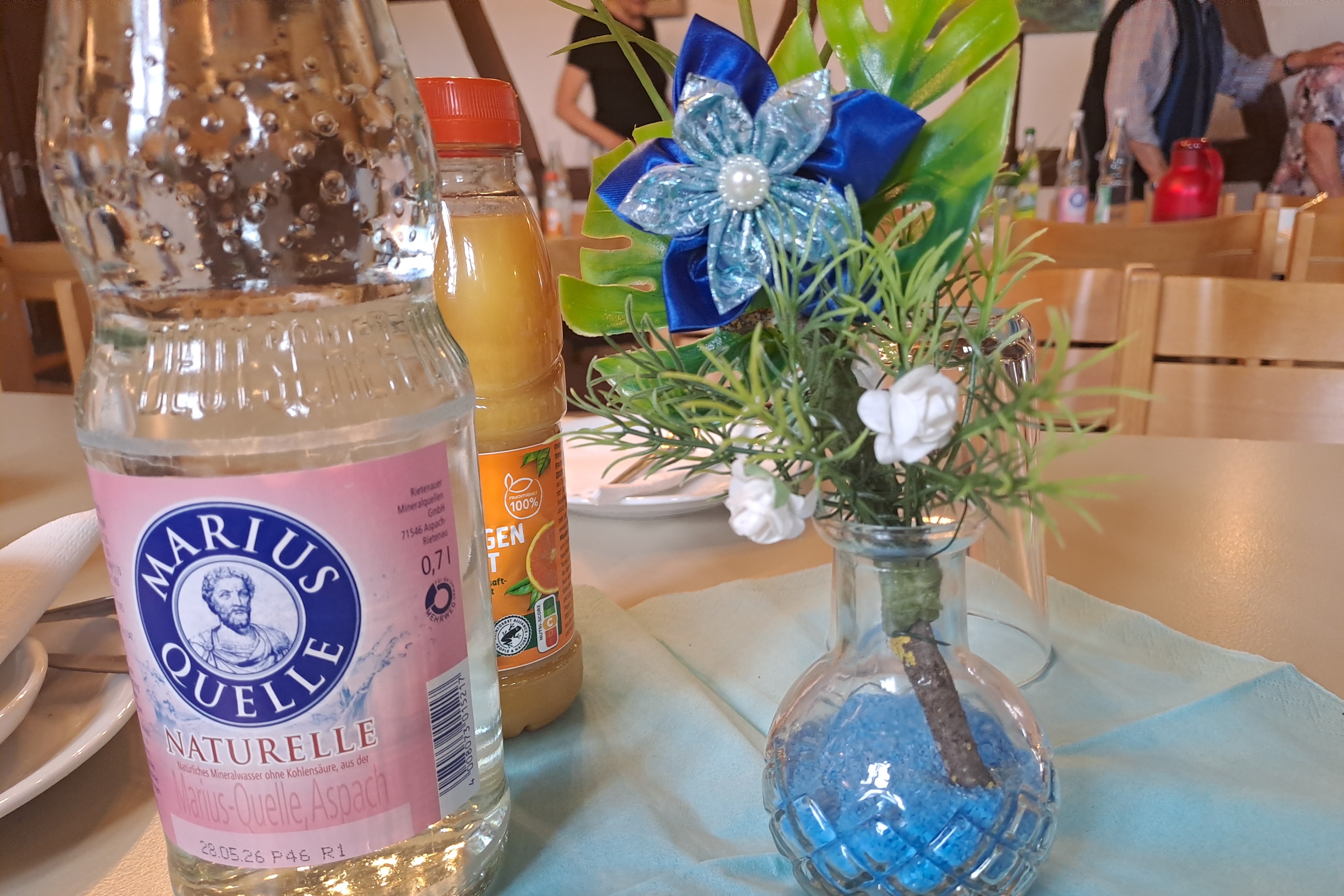 Auf dem Bild sieht man einen Tisch eine Vase mit einer blauen Blume aus Satin und Getr&auml;nken.