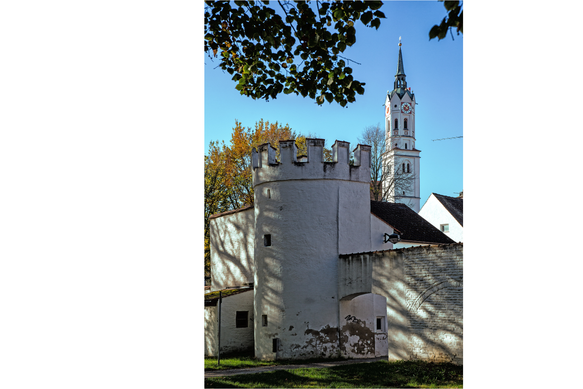 Das Bild zeigt einen Ausschnitt der Stadt Schrobenhausen bei strahlend blauen Himmel. Im Vordergrund ist die Stadtmauer abgebildet. Hinter der Stadtmauer kann man die Spitze eines Kirchturms sehen.