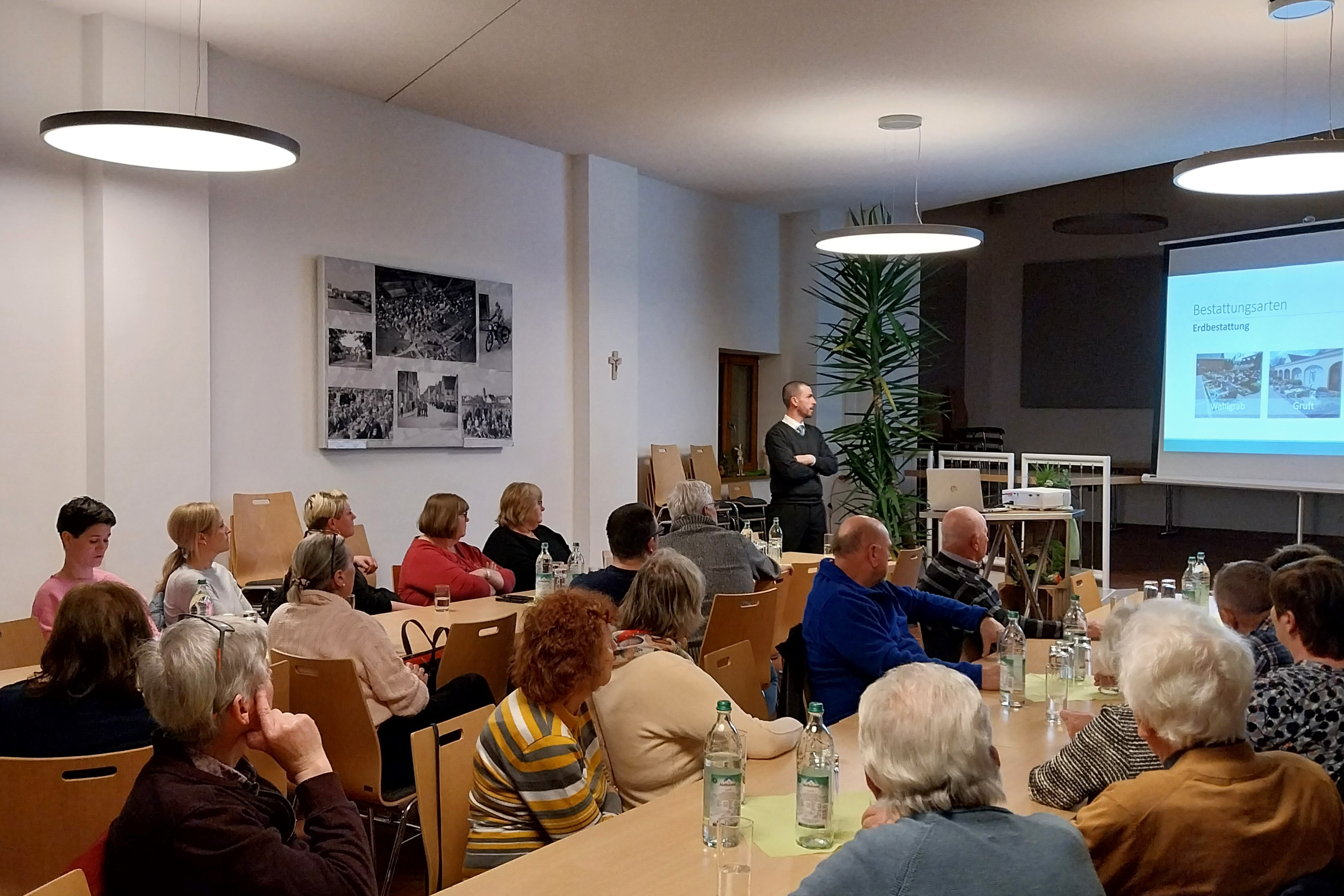 Gäste sitzen an Tischen im Vortragsraum und blicken auf die Präsentation auf der Leinwand.