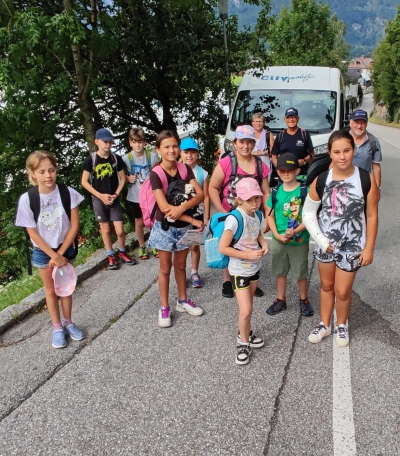 Auf dem Bild sind 9 Kinder und 3 Erwachsene als lockere Gruppe zu sehen. Im Hintergrund ist der weiße City Bus der Stadt.