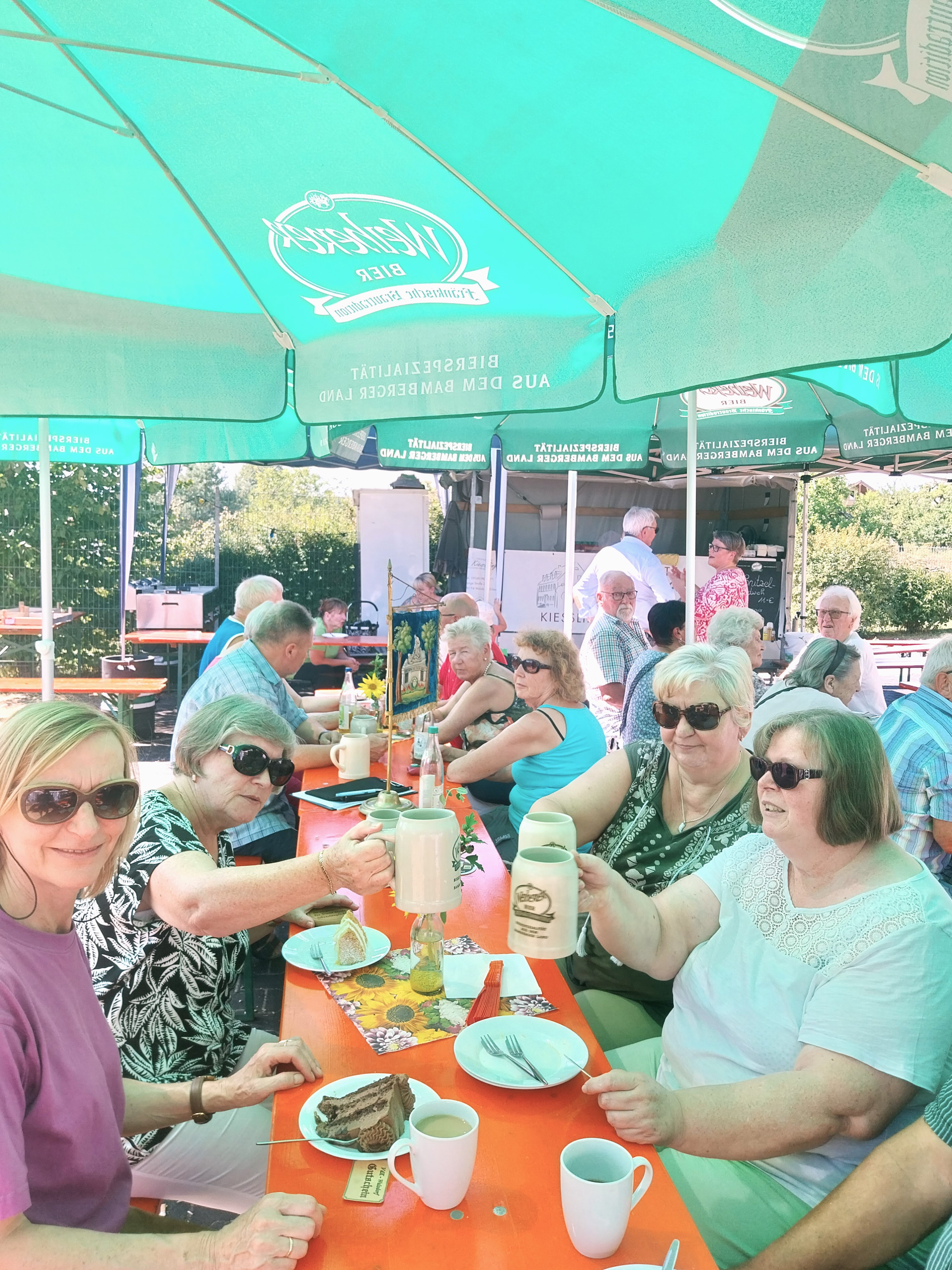 Gäste sitzen bei strahlendem Sonnenschein zum Kaffeekränzchen an Biertischen und unterhalten sich.